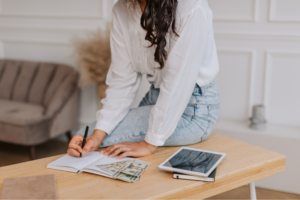 A woman sitting on top of a desk, writing in a notebook, with dollars next to her.