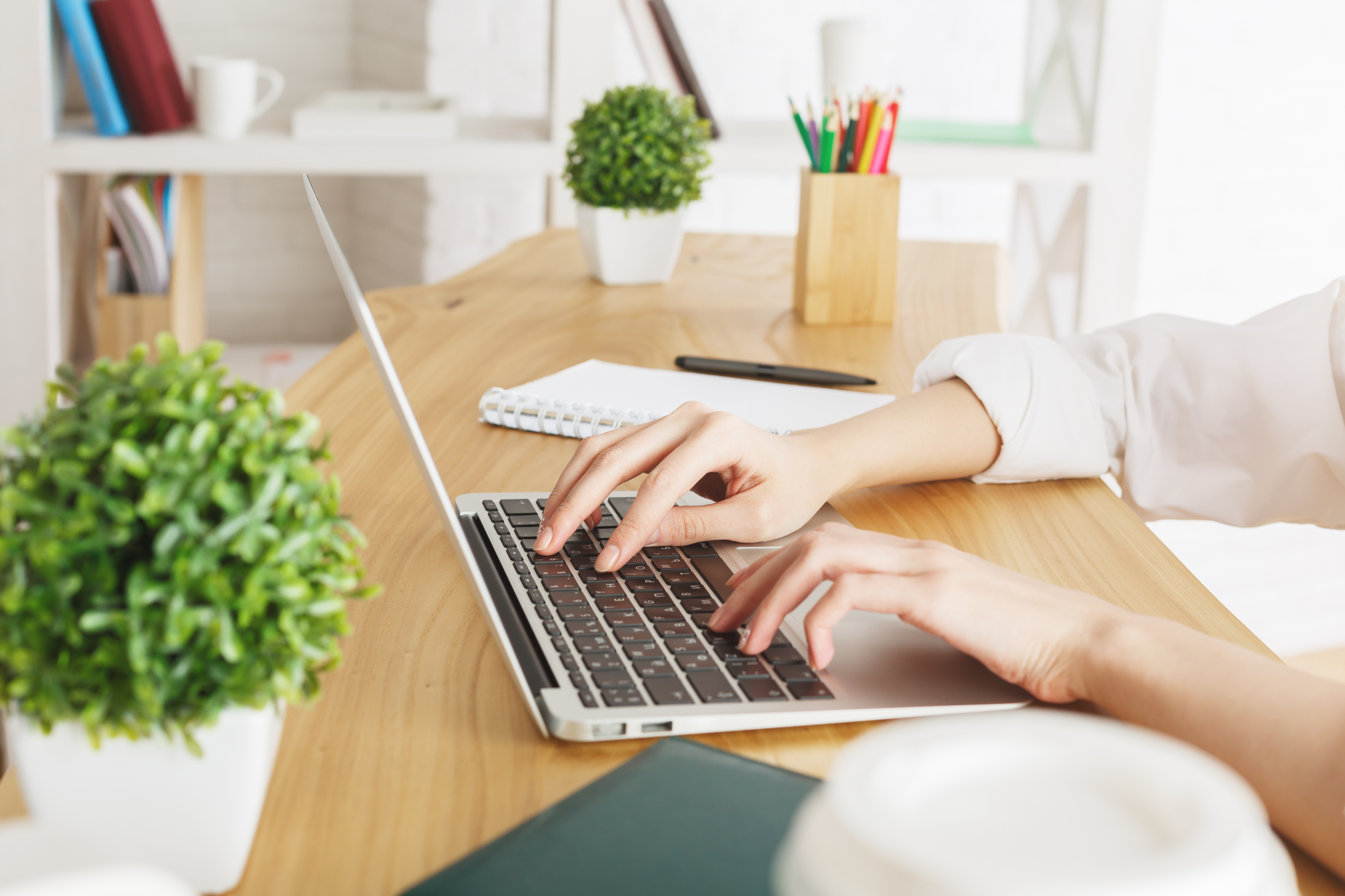 A person typing on a laptop, with a notebook and pen next to them on the desk.